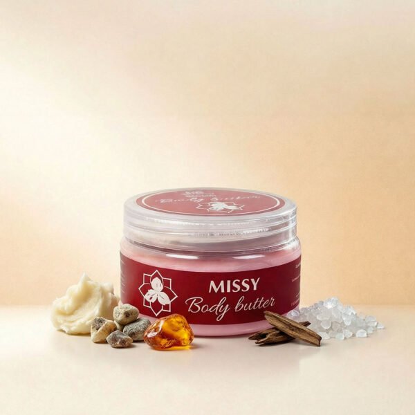 صورة إعلانية تعرض أفضل زبدة الجسم ميسي Missy Body Butter من ناتشرال كير عمان، تبرز العبوة الدائرية الشفافة بملصقها العنابي الفاخر محاطة بمكوناتها الطبيعية من أحجار المسك، العنبر، أخشاب العود، أملاح البحر الميت، وزبدة الشيا الخام على سطح زجاجي عاكس.
