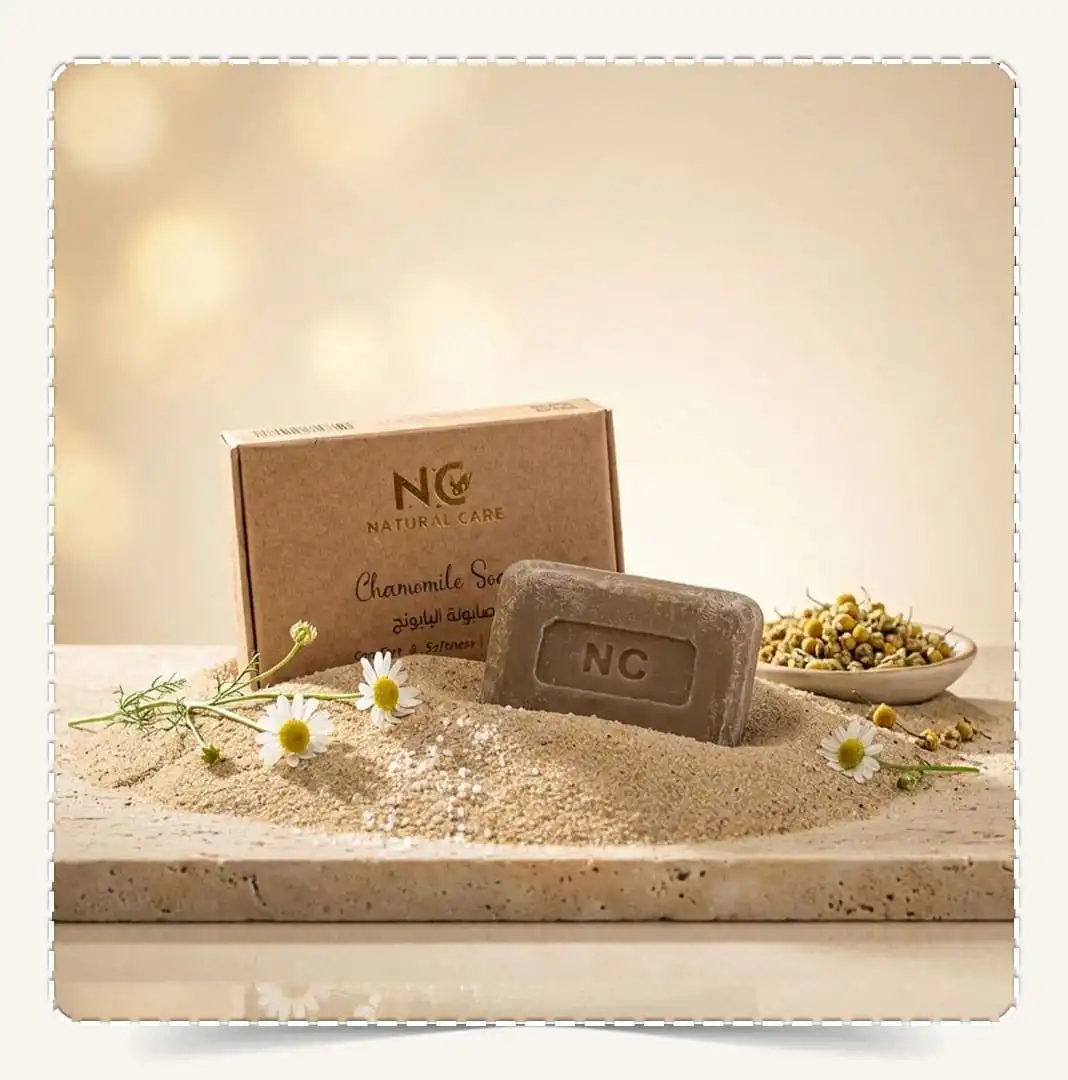 صابونة البابونج | Chamomile Soap من ناتشورال كير عمان Natural Care Oman — علبة كرافت مع صابونة NC مدفونة جزئيًا في الرمل وزهور بابونج وخلفية ذهبية ناعمة