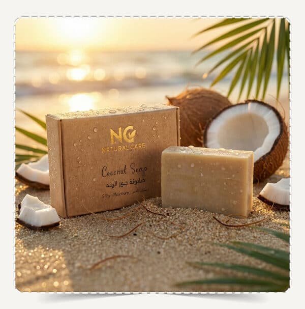 صابونة جوز الهند الطبيعية | Coconut Soap من Natural Care Oman / ناتشرال كير عمان – علبة الصابون وقطعة الصابون على رمال الشاطئ مع جوز هند وقطرات ماء وأجواء غروب