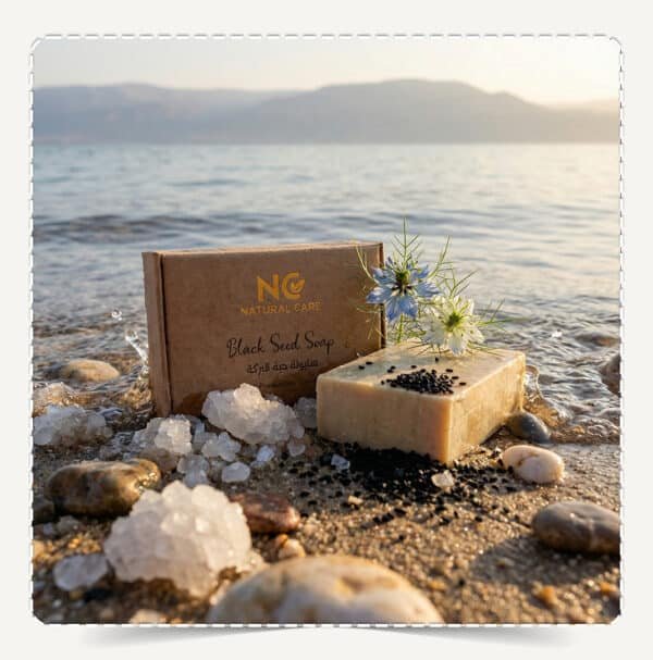 صابونة حبة البركة الطبيعية | Black Seed Soap من Natural Care Oman / ناتشرال كير عمان — المنتج على شاطئ مع مياه وحصى وبلورات ملح وبذور حبة البركة وزهرة لإيحاء نقاء وانتعاش طبيعي