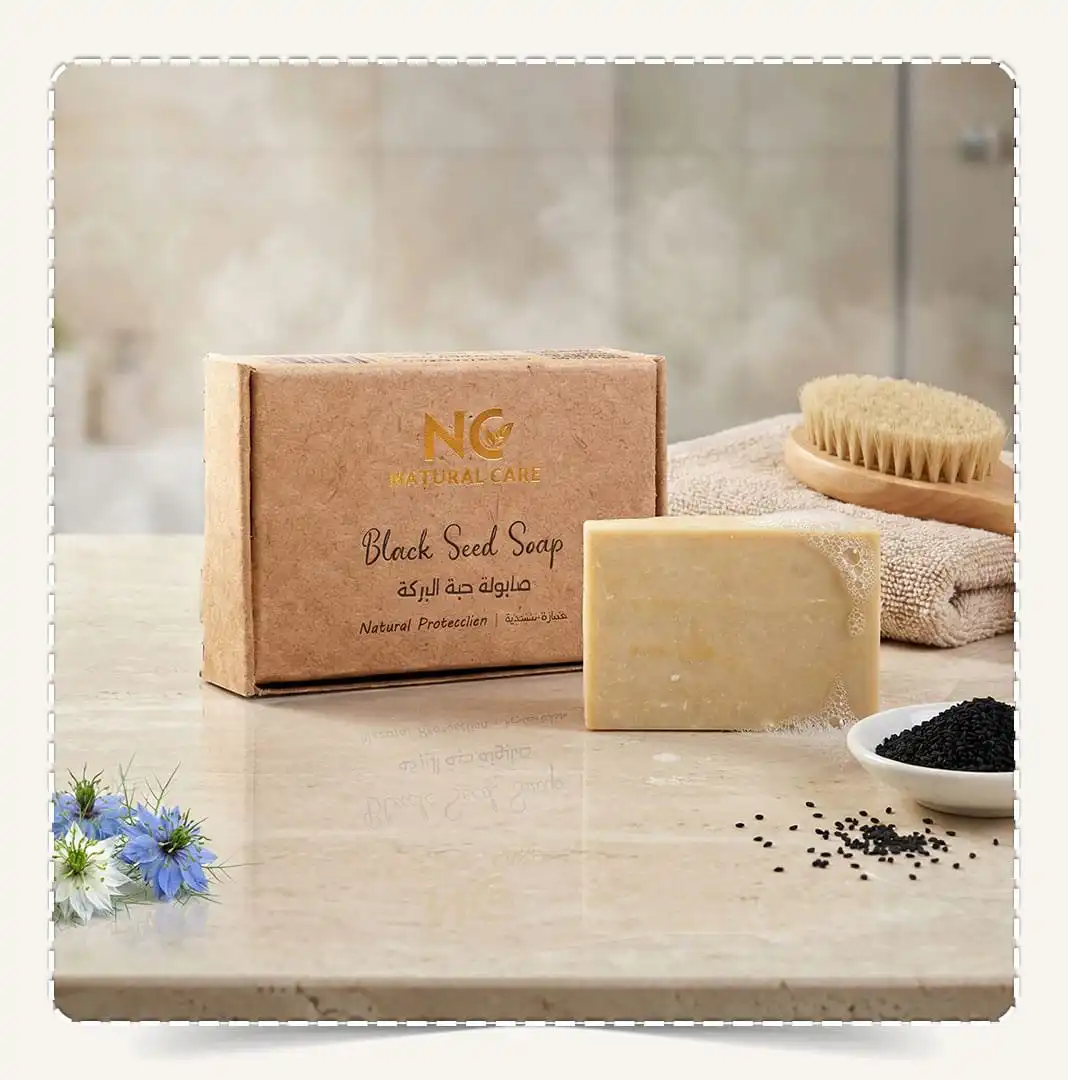 صابونة حبة البركة الطبيعية | Black Seed Soap من Natural Care Oman / ناتشرال كير عمان — علبة المنتج وقطعة الصابون على سطح رخامي مع فرشاة خشبية ومنشفة وبذور حبة البركة لإيحاء تنظيف عميق وروتين عناية يومي