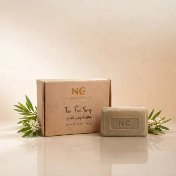 صندوق وقطعة صابونة شجرة الشاي الطبيعية | Tea Tree Soap من ناتشرال كير عمان، بجوار أوراق شجرة الشاي الخضراء النضرة وزجاجة زيت شجرة الشاي العطري لتعكس النقاء والتوازن.