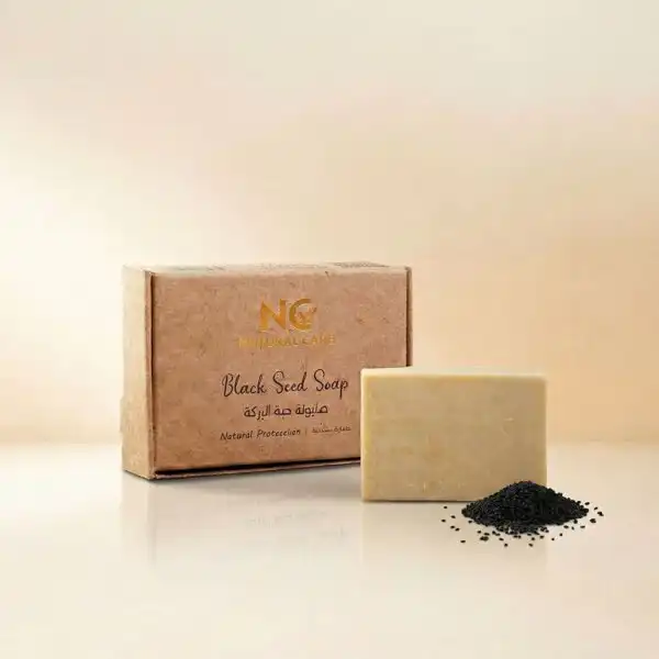 صندوق وقطعة صابونة حبة البركة الطبيعية - Black Seed Soap من ناتشرال كير عمان، بجوار كومة صغيرة من بذور حبة البركة السوداء النقية لتعكس الحماية الطبيعية والتنقية العميقة.
