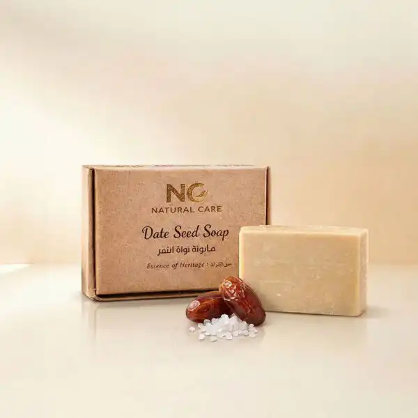 صندوق وقطعة صابونة نواة التمر الطبيعية - Date Seed Soap من ناتشرال كير عمان، بجوار حبات التمر الطازجة وأملاح البحر الميت لتعكس عبق التراث والعناية العضوية الأصيلة.