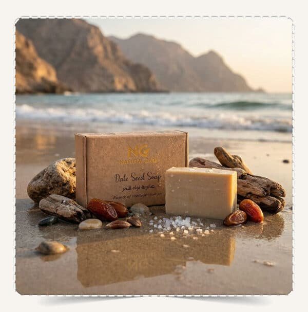 صابونة نواة التمر الطبيعية | Date Seed Soap من Natural Care Oman / ناتشرال كير عمان – المنتج على رمل الشاطئ أمام البحر مع صخور وتمر ونوى لإيحاء طبيعي
