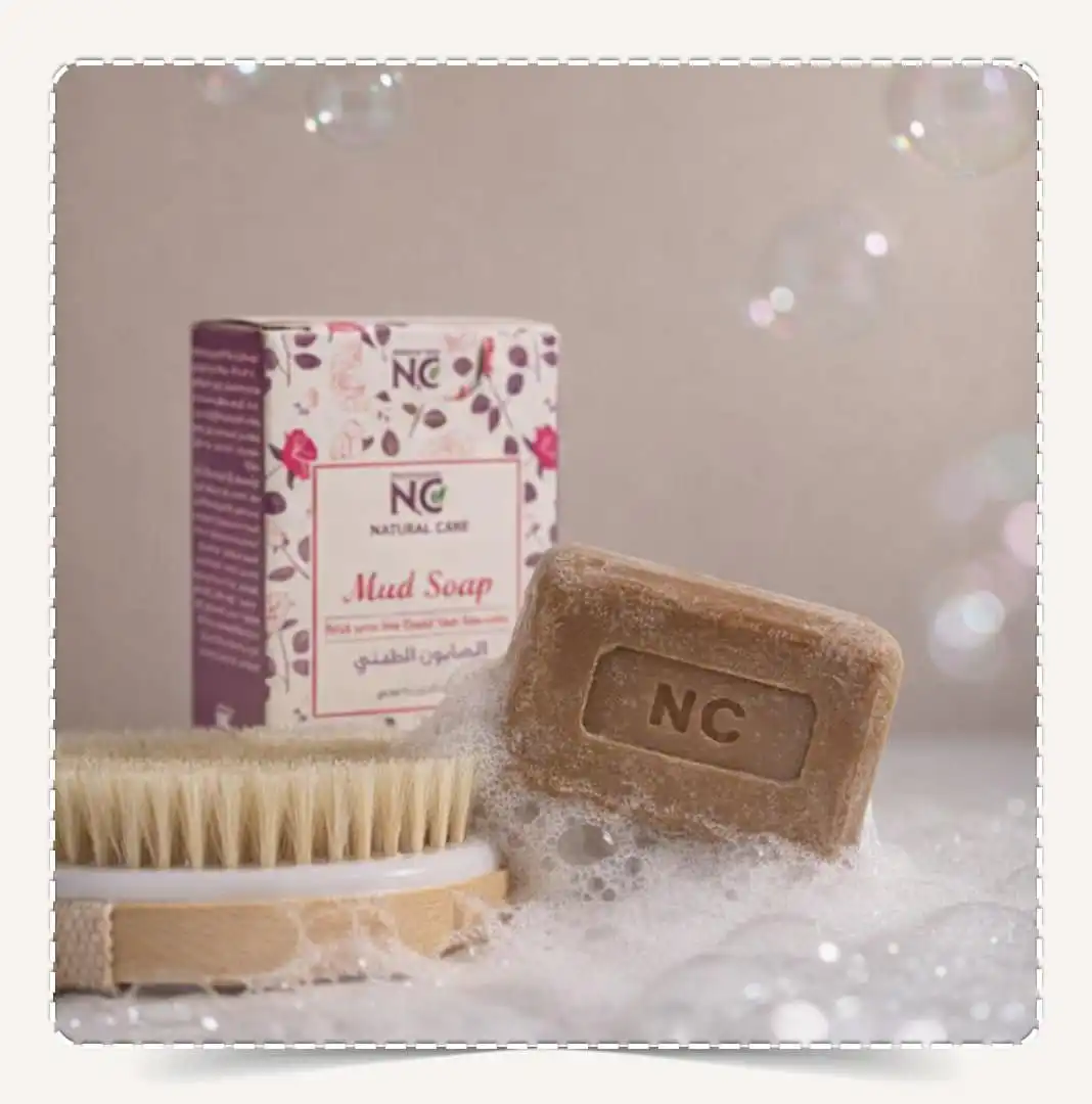 صابون الطين بأملاح البحر الميت | Mud Soap من ناتشورال كير عمان Natural Care Oman — قالب صابون طيني داخل رغوة حمام مع فرشاة والعبوة بالخلف
