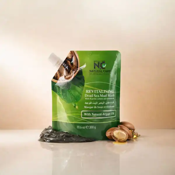 عبوة قناع طين البحر الميت للوجه بزيت الأرجان Revitalising Dead Sea Mud Mask من ناتشرال كير عمان، موضوعة بجوار طين البحر الميت الخام وثمار وزيت الأرجان الذهبي.