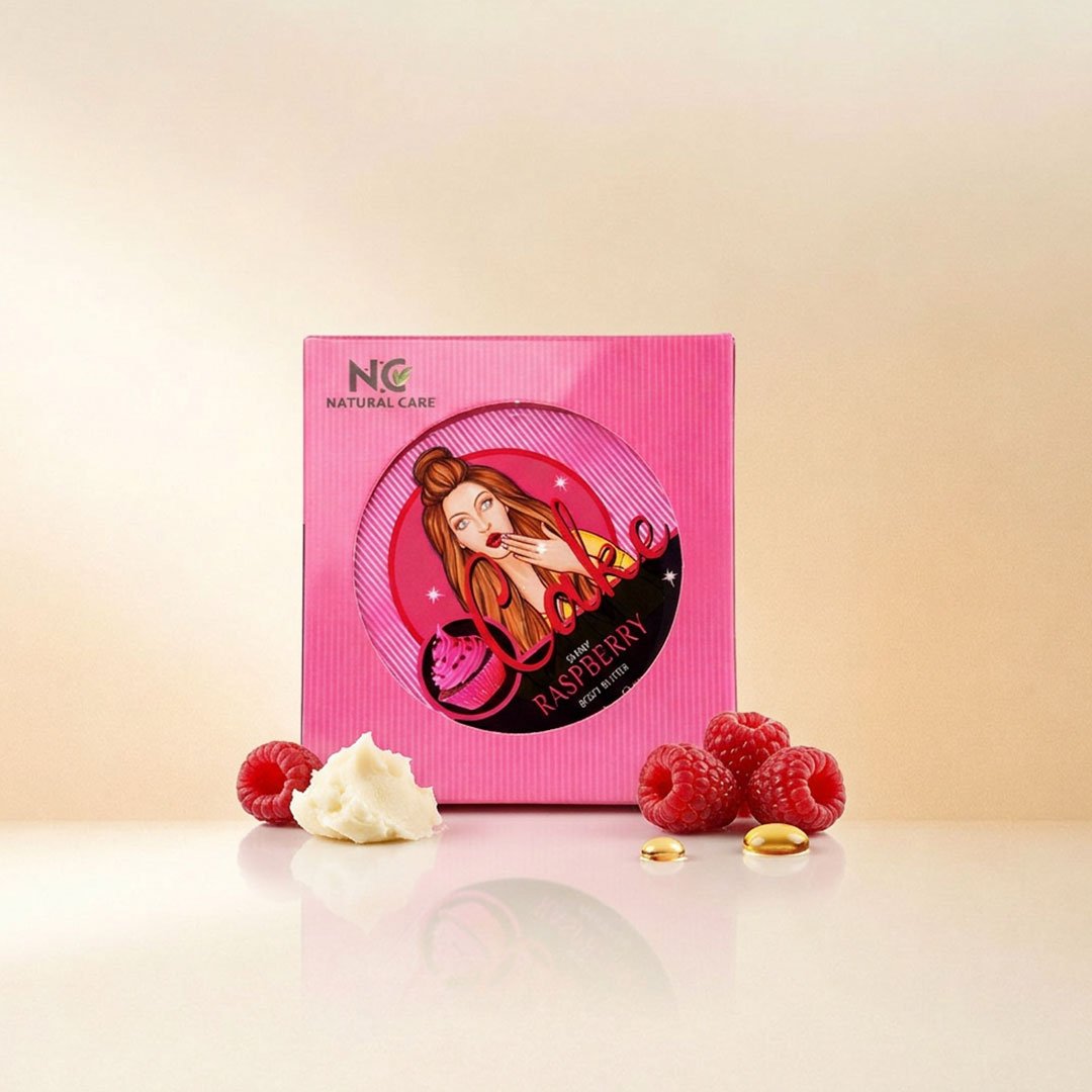 صورة تعرض أفضل زبدة الجسم الطبيعية راسبيري Raspberry Natural من ناتشرال كير عمان، يبرز صندوق التغليف الوردي الجذاب محاطاً بحبات التوت الأحمر الطازجة، قطعة من الزبدة الخام، وقطرات الزيوت الطبيعية على سطح زجاجي عاكس.